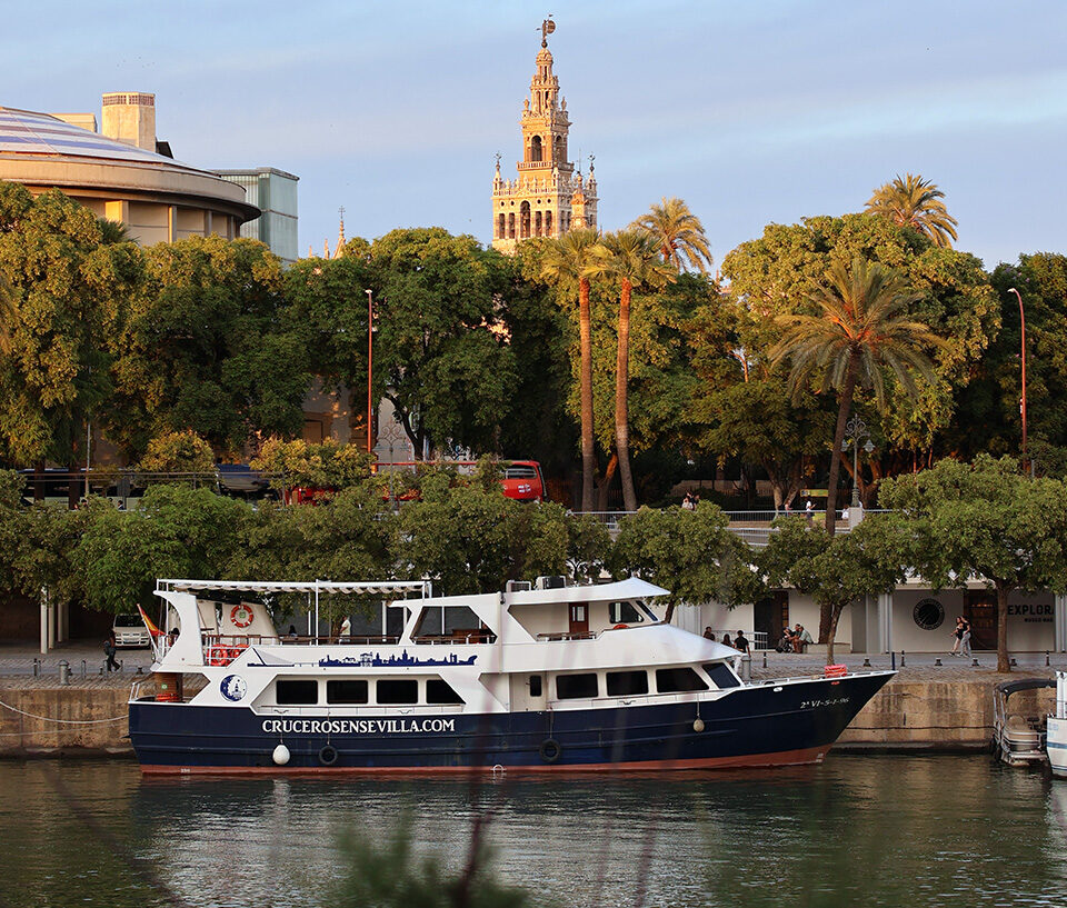 Barco turístico atracado en el río Guadalquivir en Sevilla, con la Giralda al fondo entre las palmeras al atardecer.
